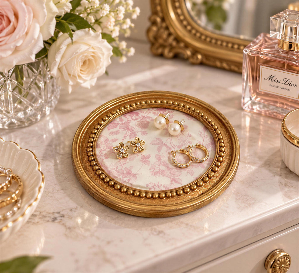 A round gold tray with a floral pattern holds a pair of earrings and a perfume bottle on a white marble countertop.