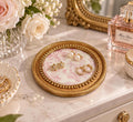 A round gold tray with a floral pattern holds a pair of earrings and a perfume bottle on a white marble countertop.