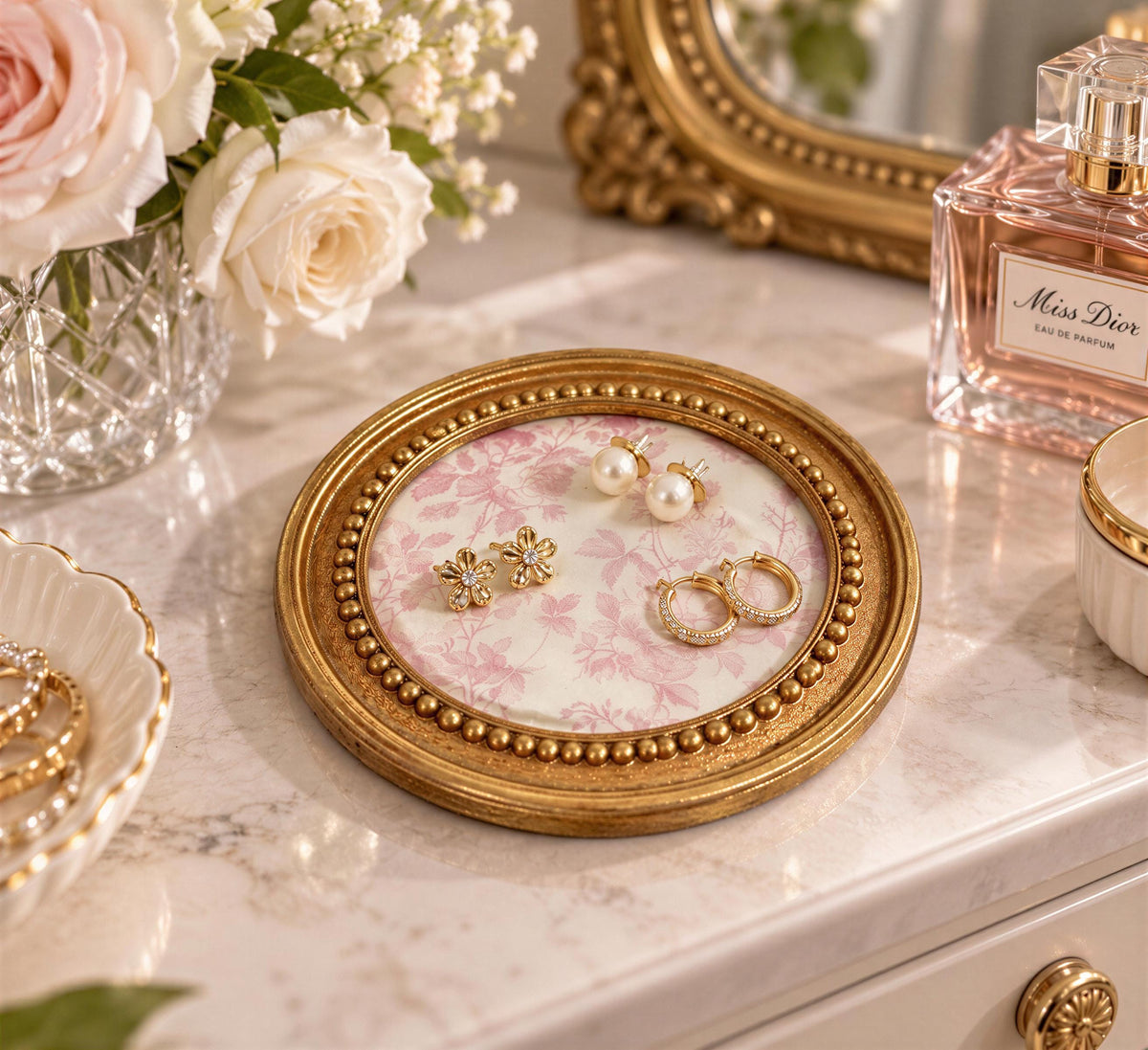 A round gold tray with a floral pattern holds a pair of earrings and a perfume bottle on a white marble countertop.