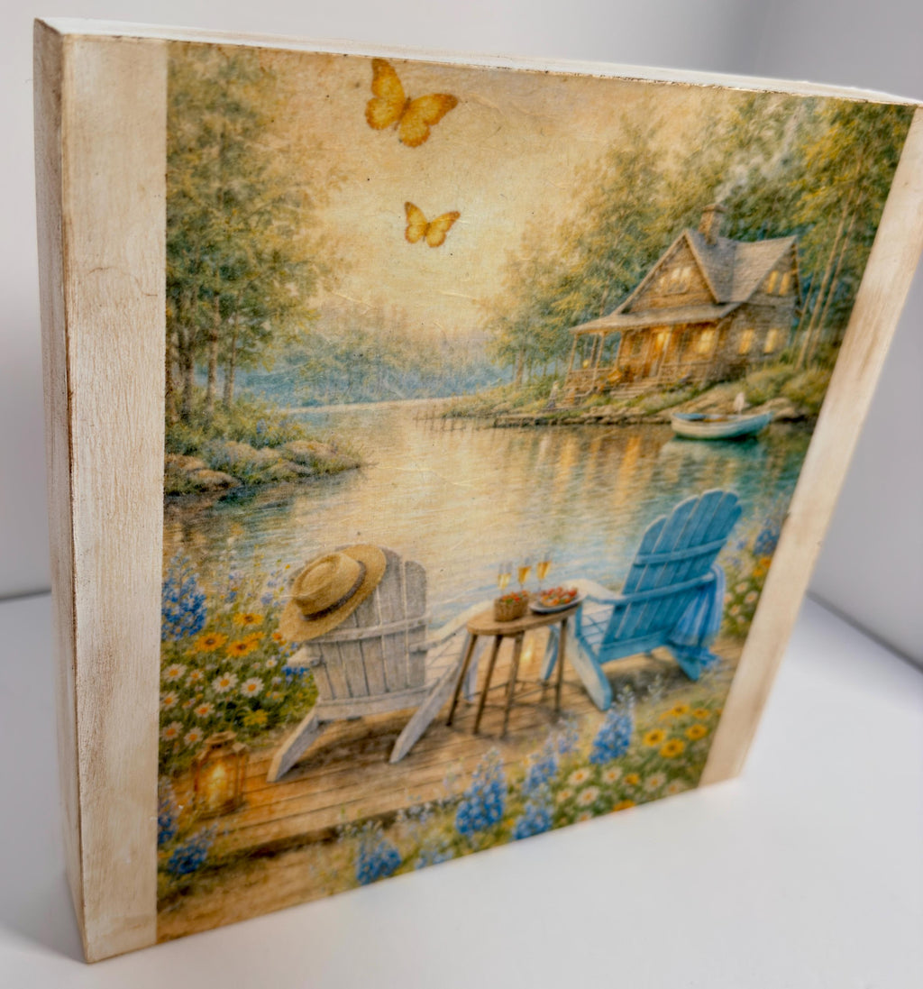 A painting depicting a serene lakeside scene with a cabin, two chairs, and a table, surrounded by flowers and butterflies.