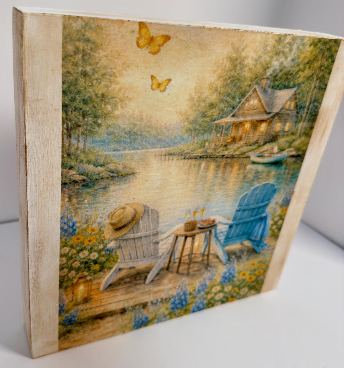 A painting depicting a serene lakeside scene with a cabin, two chairs, and a table, surrounded by flowers and butterflies.
