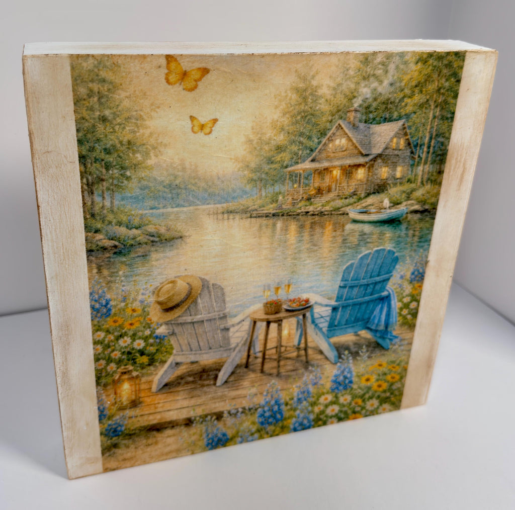 A wooden cabin with a porch overlooking a lake, surrounded by trees and flowers. Two Adirondack chairs are placed on a wooden deck, with a table and a vase of flowers nearby. Butterflies can be seen in the sky, and a boat is visible on the lake.