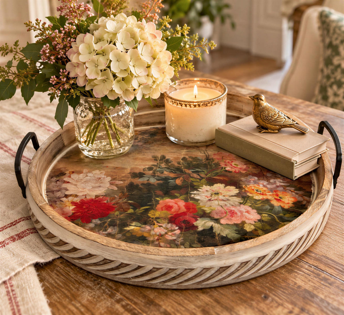 A wooden tray with a floral design sits on a wooden table, accompanied by a candle, a book, and a small bird figurine.
