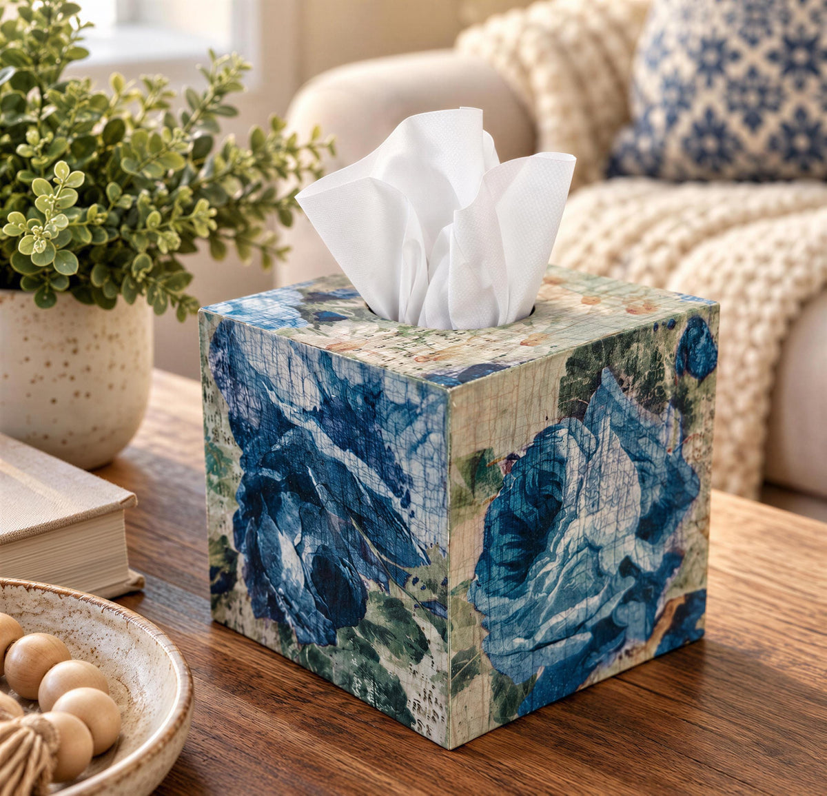 A wooden tissue box with a floral pattern sits on a wooden table, surrounded by various decorative items such as a potted plant, a bowl, and a book.