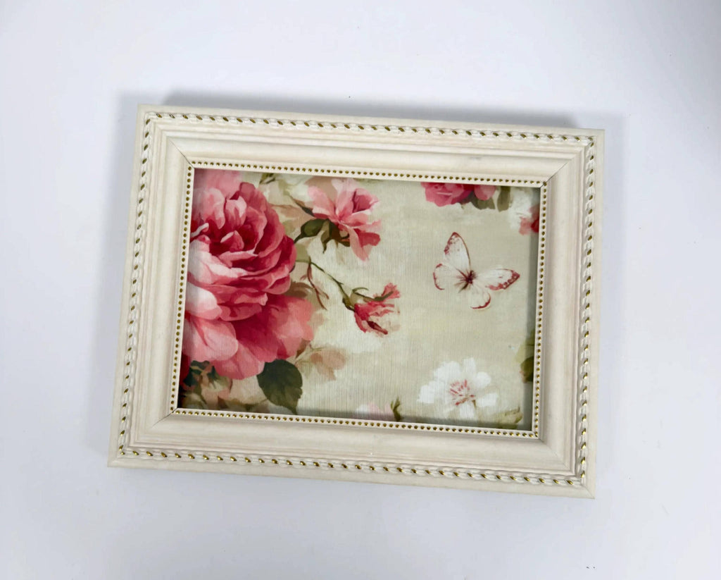 A framed picture with a floral design, featuring a large pink rose and a butterfly.
