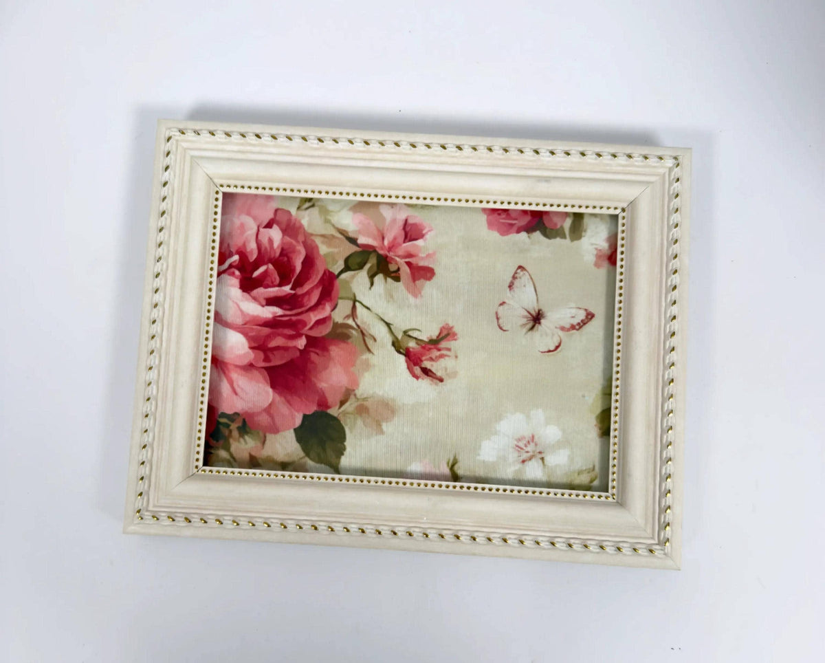 A framed picture with a floral design, featuring a large pink rose and a butterfly.