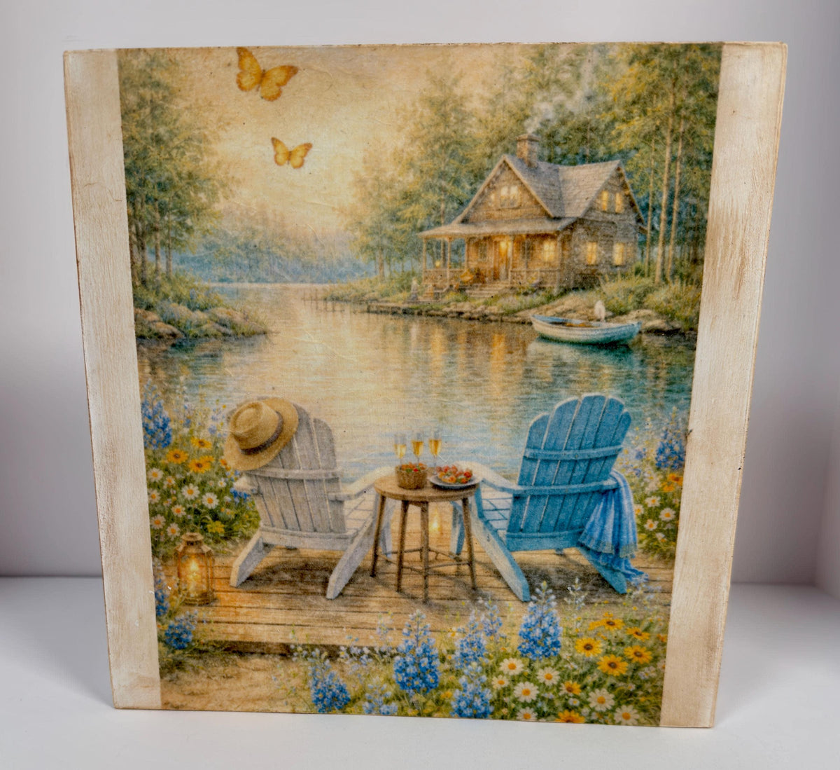 A painting depicting a serene lakeside scene with a cabin, two blue Adirondack chairs, and a table with a drink set up, surrounded by flowers and butterflies.