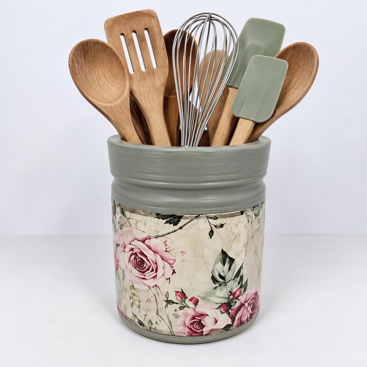 A gray ceramic jar with a floral pattern, containing various kitchen utensils such as wooden spoons, a whisk, and a spatula.