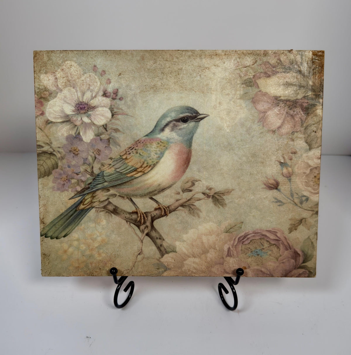 a square-shaped piece of art with a painting of a bird perched on a branch, surrounded by flowers and foliage.