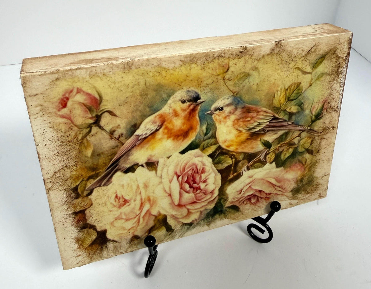 a wooden block with a painting of two birds perched on flowers, including roses.