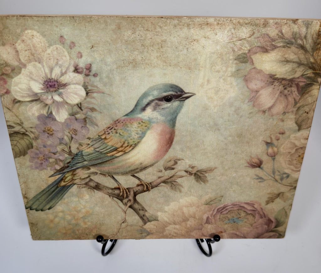 a square-shaped piece of art with a painting of a colorful bird perched on a branch, surrounded by flowers and foliage.