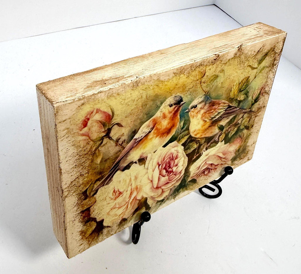 a wooden block with a painting of two birds perched on flowers, including roses.