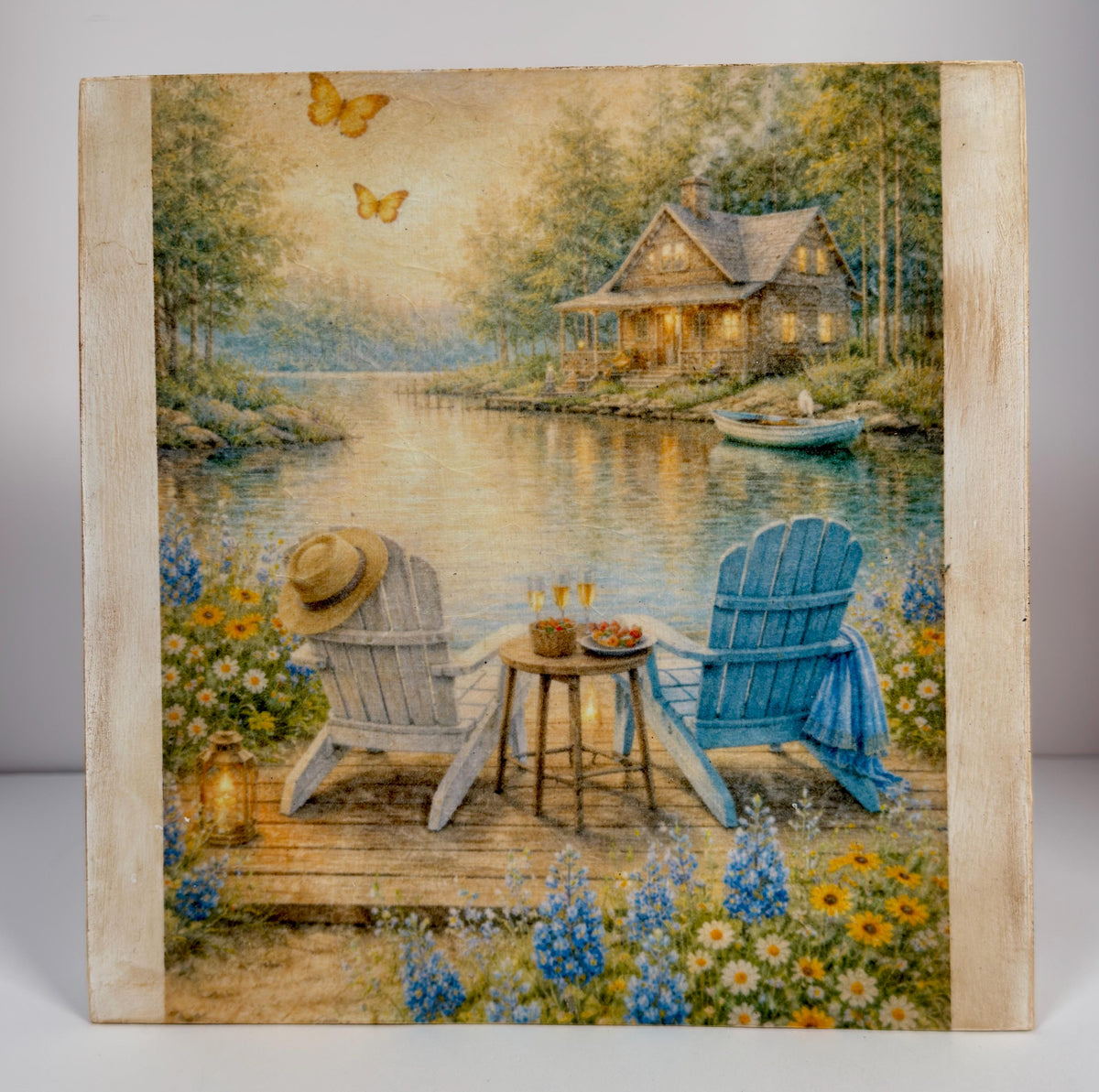 A painting depicting a serene lakeside scene with a cabin, two Adirondack chairs, and a table with a vase of flowers. Butterflies are flying in the sky, and a boat is on the water.
