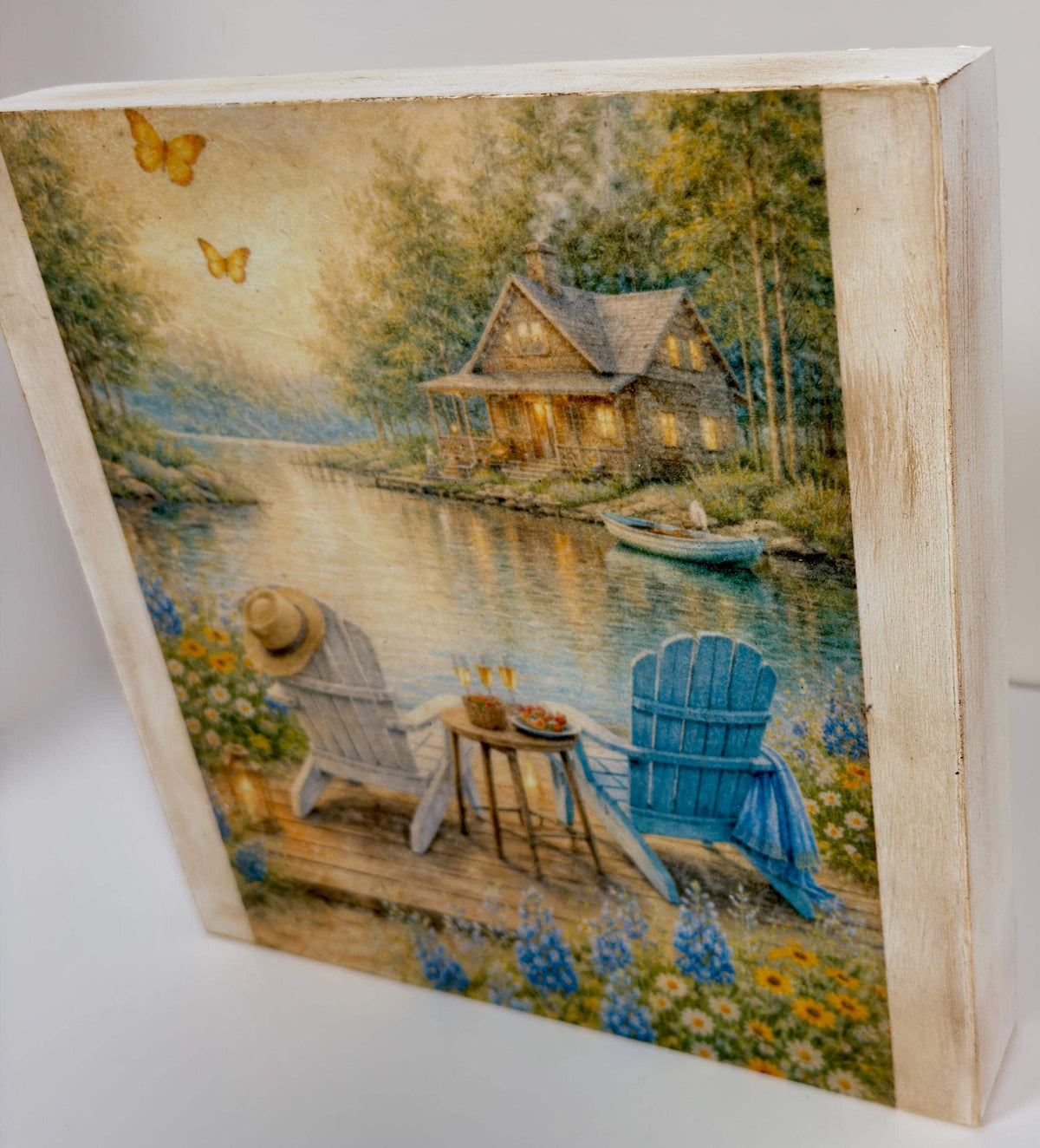 A painting depicting a serene lakeside scene with a cabin, two blue chairs, and a boat on the water.