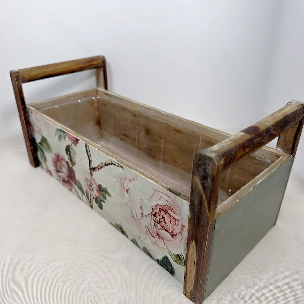 A wooden box with a floral pattern on the front, featuring a handle on each side.