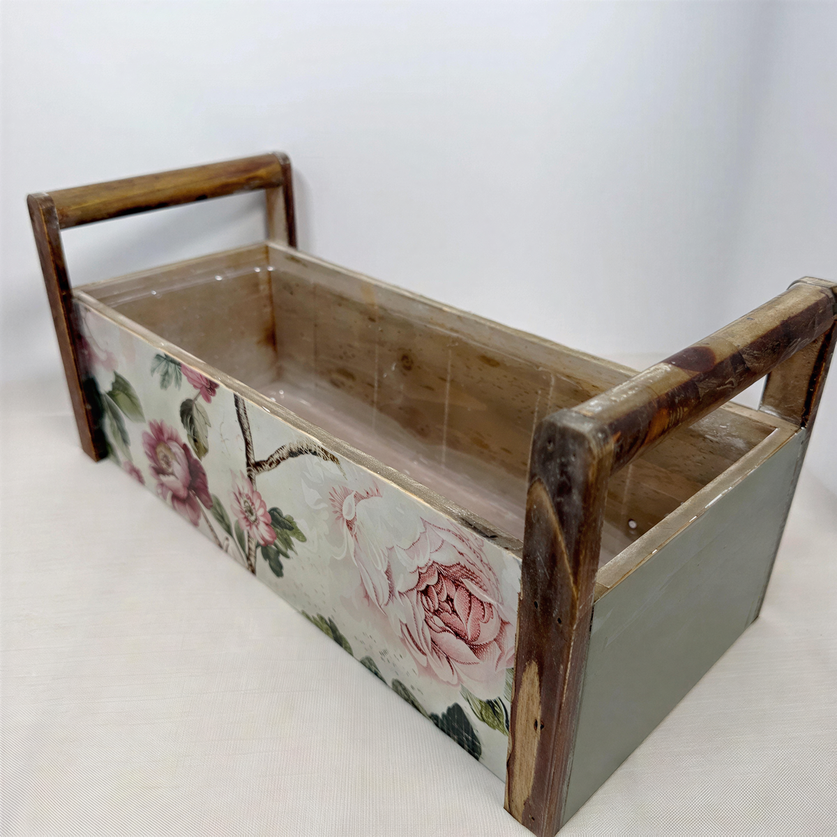 A wooden box with a floral pattern on the front, featuring a handle on each side.