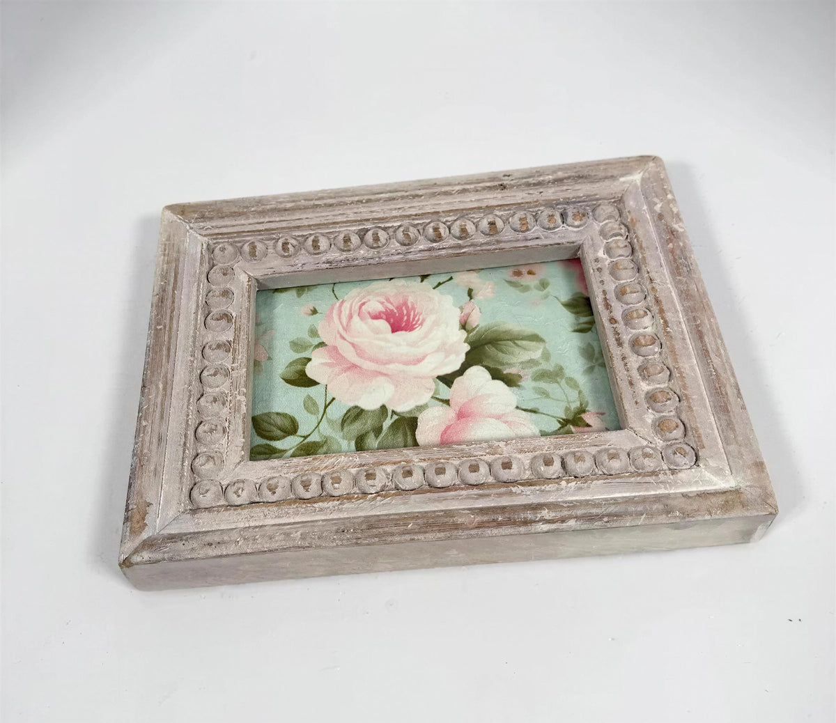 A small, square picture frame with a floral design inside, placed on a white surface.