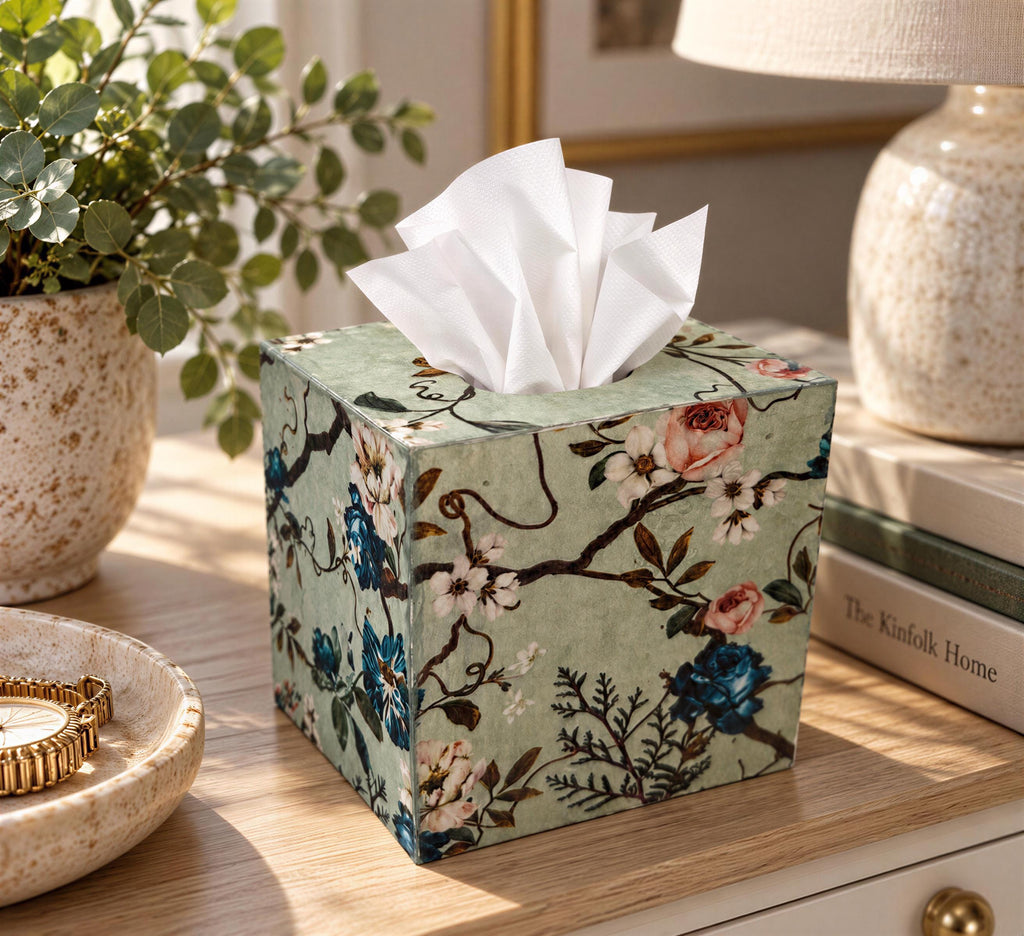 A decorative tissue box with a floral pattern sits on a wooden surface, surrounded by various objects such as a potted plant, a lamp, and books.