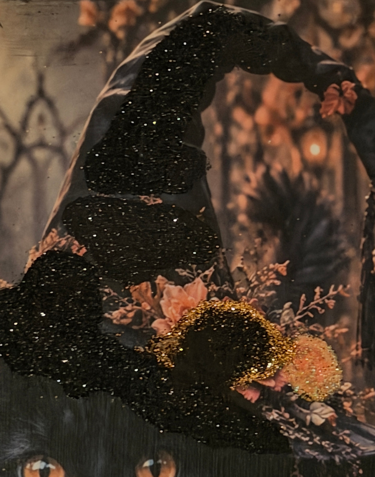 A close-up of a black witch's hat adorned with gold glitter and flowers, with a blurred background that appears to be a garden or forest.
