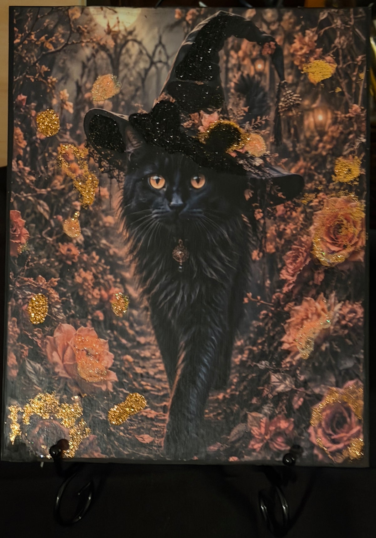 A black cat wearing a witch's hat, surrounded by a colorful autumnal scene with flowers and foliage.