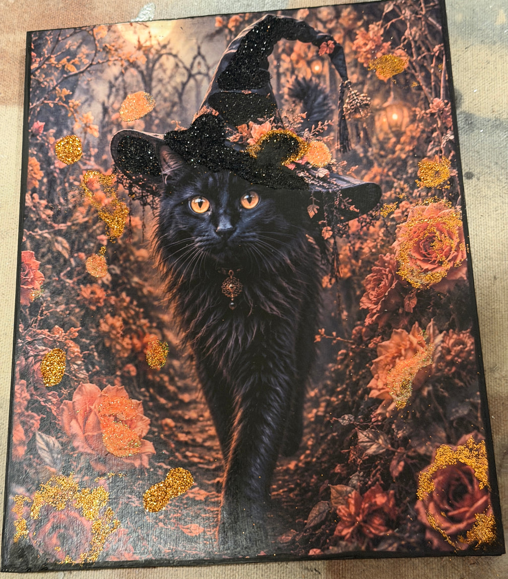 A black cat wearing a witch's hat, surrounded by autumn foliage and flowers, creating a whimsical and festive scene.