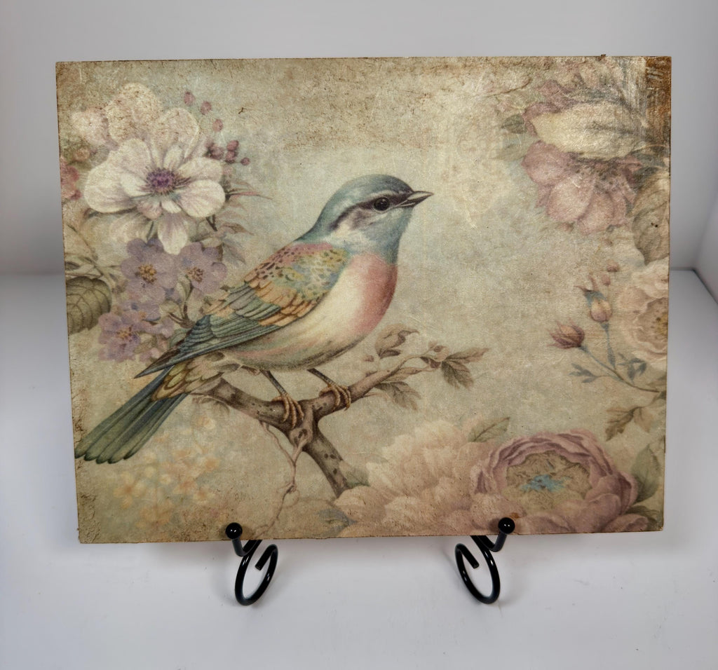 a square-shaped piece of art with a painting of a bird perched on a branch, surrounded by flowers and foliage.