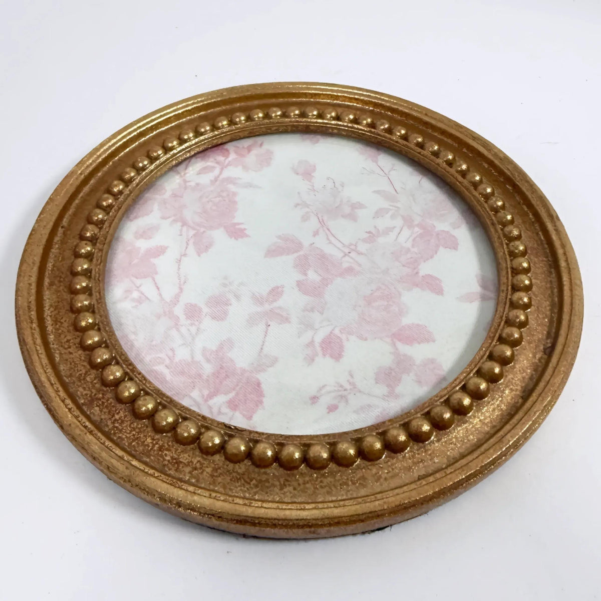 A round gold-colored picture frame with a floral pattern inside, placed on a white surface.