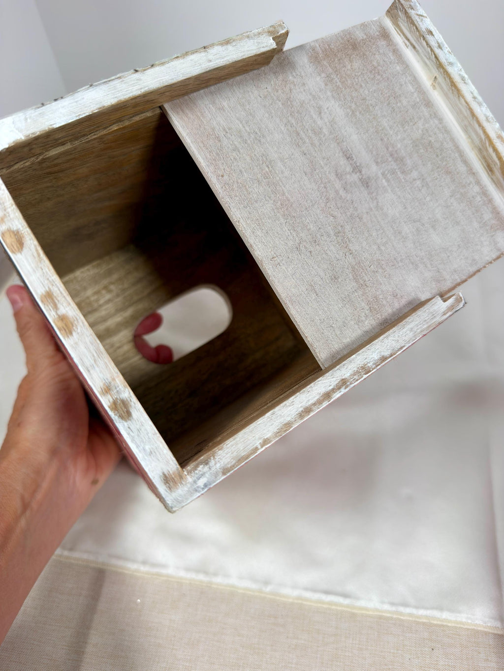 A hand is holding a wooden box with a white interior, revealing a small red object inside.