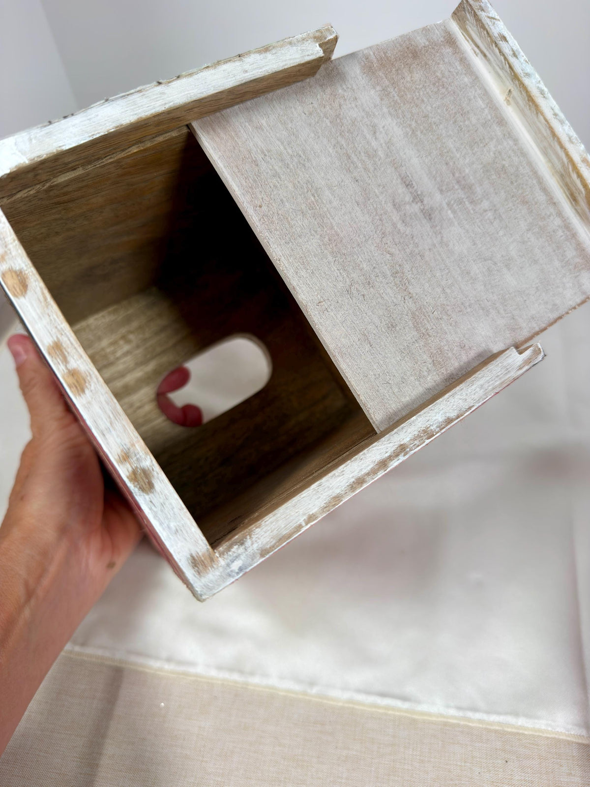 A hand is holding a wooden box with a white interior, revealing a small red object inside.