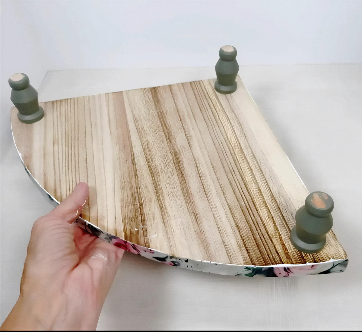 A hand is holding a wooden tray with a floral pattern on it, which appears to be made of wood and has three green wooden legs attached to it.