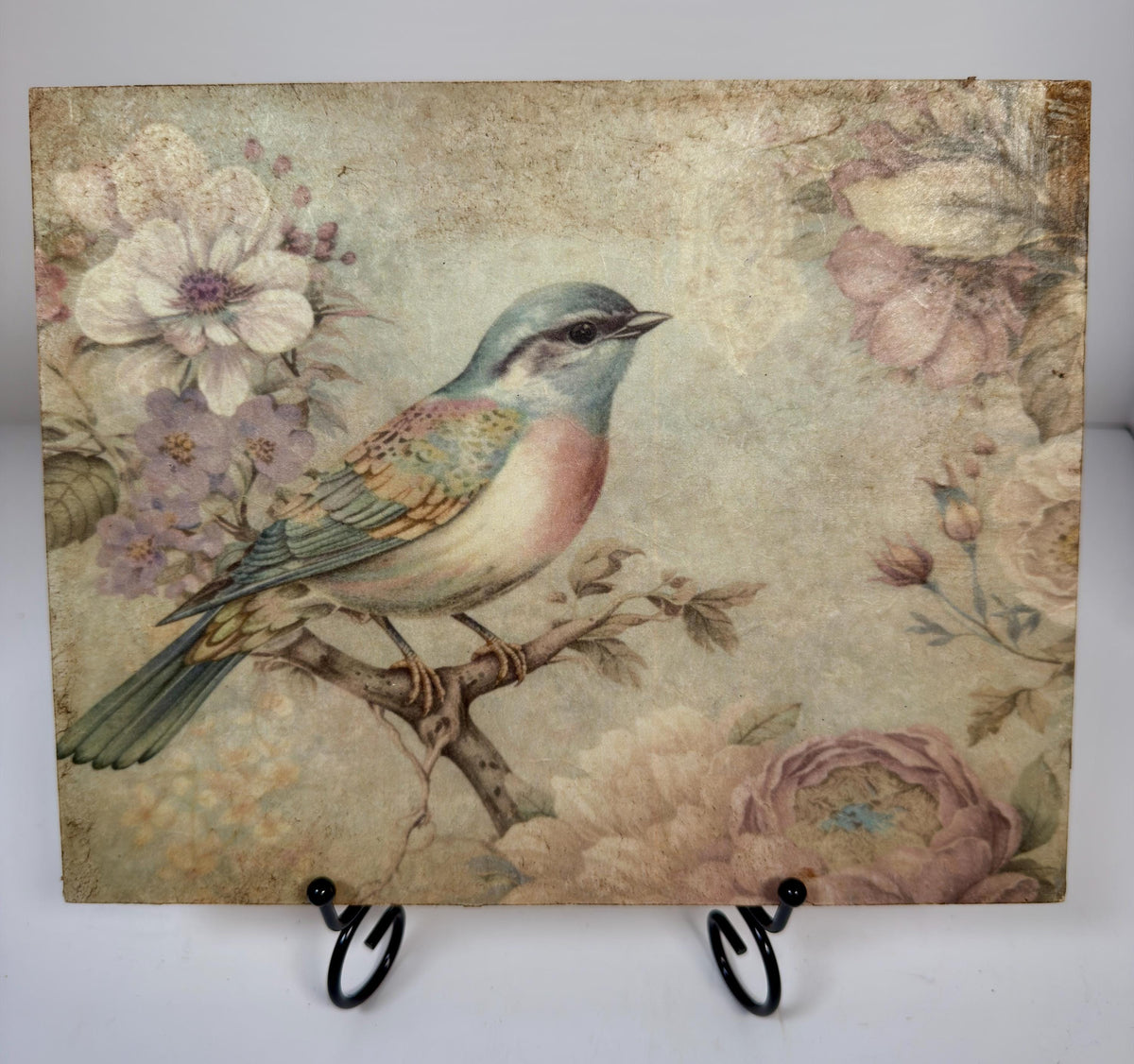 a square-shaped piece of art with a painting of a bird perched on a branch, surrounded by flowers and foliage.