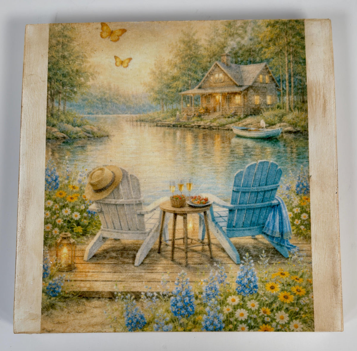 A painting depicting a serene lakeside scene with a cabin, two Adirondack chairs, and a table with drinks, surrounded by flowers and butterflies.