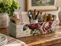 A wooden crate with a floral design, containing various kitchen utensils such as knives, spoons, and a vase.